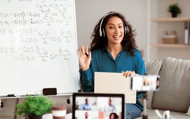 Distance Education. Portrait of teacher in wireless headset having virtual meeting with students on laptop, sitting at desk, waving hand to webcam shooting video training on smartphone using tripod