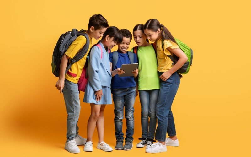 Pupils, Technology And Learning Concept. Smiling cute boy holding digital tablet, showing group of multiracial classmates friends nice education app, studying at elementary school, yellow background