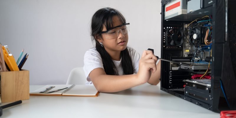 Photo of young adorable girl fixing/installing a computer hardware at the modern white working desk. Genius kid with various actions concept.