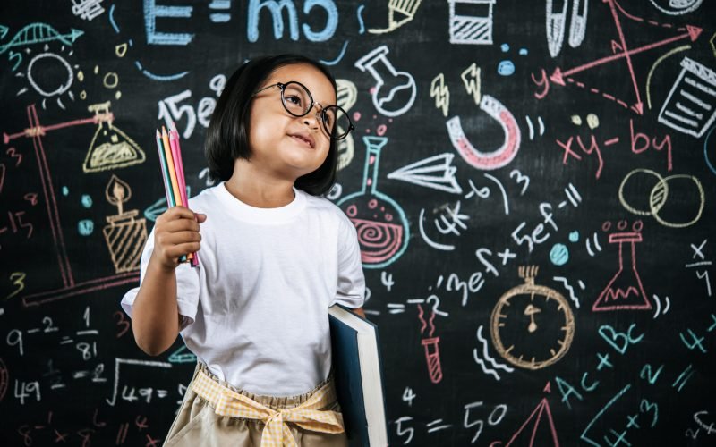 Selective focus, Little girl wearing eyeglasses holding colors pencil in hand and holding large textbook standing at front of blur blackboard in background, smile with happy