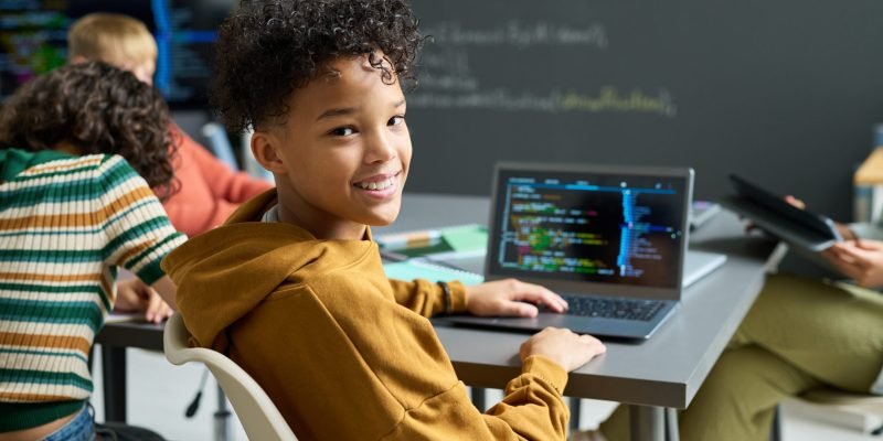 Portrait of Black teenager boy smiling and turning toward camera while coding on laptop in classroom, classmates working at desks in background, chalkboard with programming notes visible