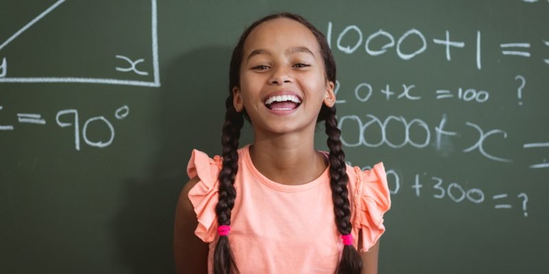 Portrait of laughing mixed race schoolgirl standing in front of chalkboard in classroom maths lesson. childhood and education at elementary school.