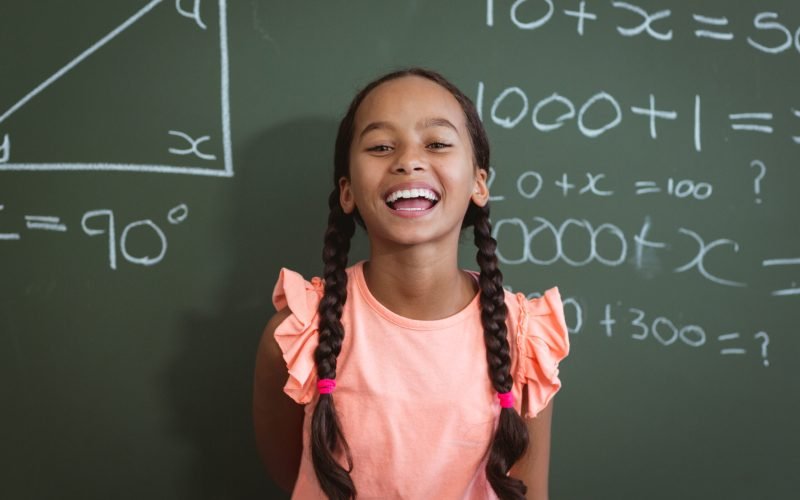 Portrait of laughing mixed race schoolgirl standing in front of chalkboard in classroom maths lesson. childhood and education at elementary school.