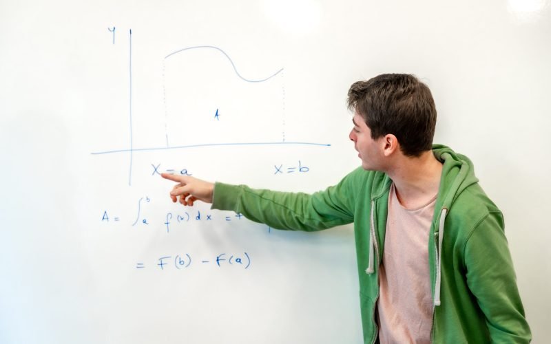 Young college student explaining a mathematical integral calculus formula, pointing at a graph on a whiteboard