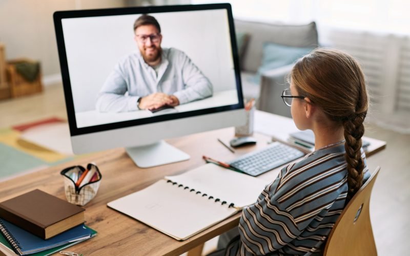 Caucasian girl sitting at desk participating in online lesson with Caucasian middle aged man on computer monitor, writing in notebook, wearing glasses, studying in home environment